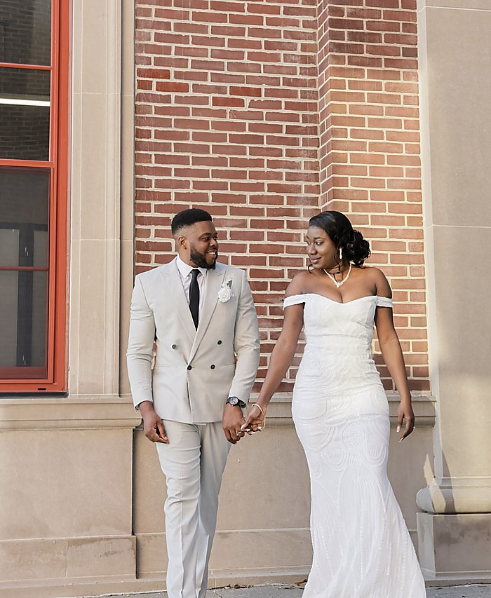 A couple in white holding hands outside a brick building: a cream suit and an off-shoulder gown