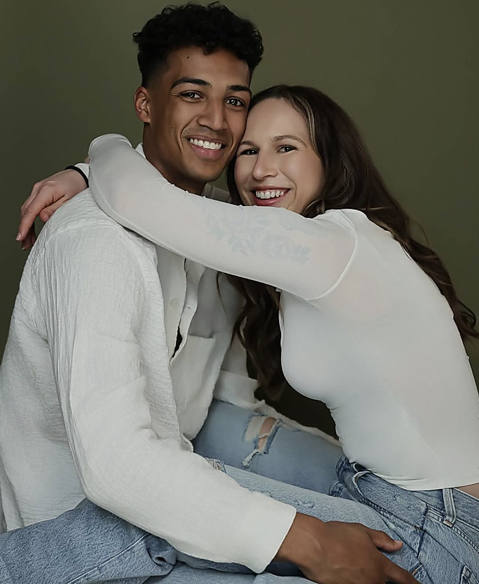 A couple on an olive backdrop, her arms around his shoulders, both smiling