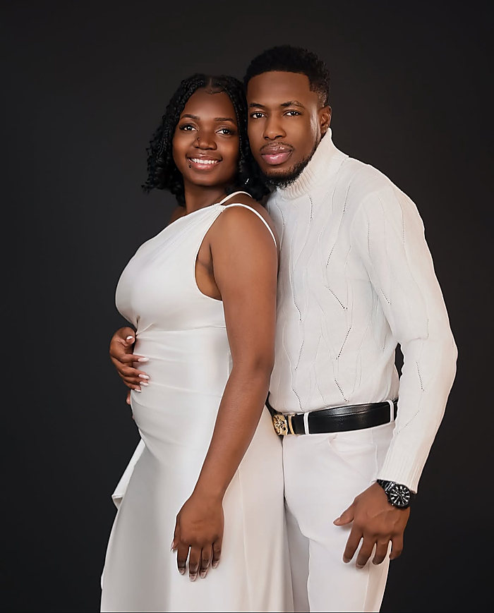 A couple in white on a dark gray backdrop, his arm around her waist