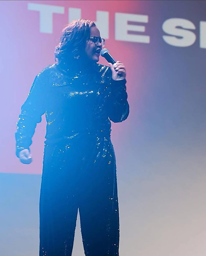 A singer on stage in a sequined jumpsuit, lit blue and pink, the screen behind