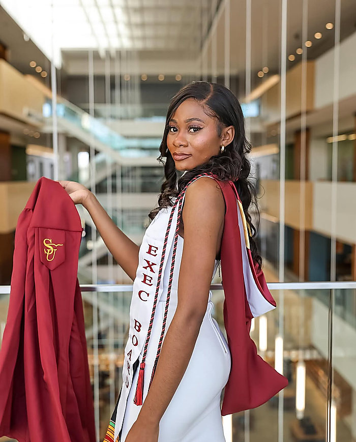 A graduate in a glass atrium, her maroon gown over one shoulder, a white dress and a stole