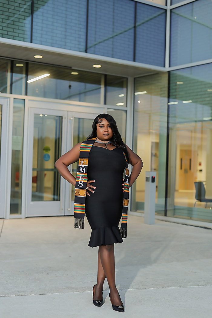 A graduate at a glass entrance: a black dress and a kente stole