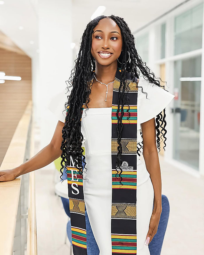 A graduate in a white dress and a kente stole in a bright hallway of glass and white walls