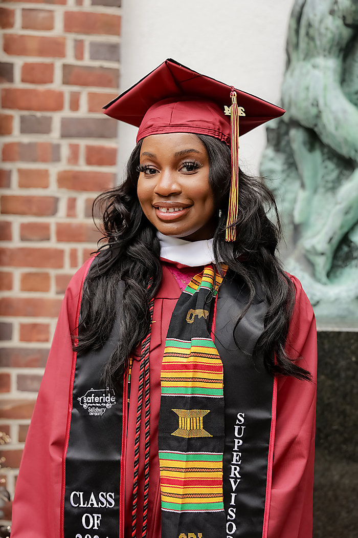 A graduate in a red cap and gown with a kente stole, beside a brick wall and a bronze statue