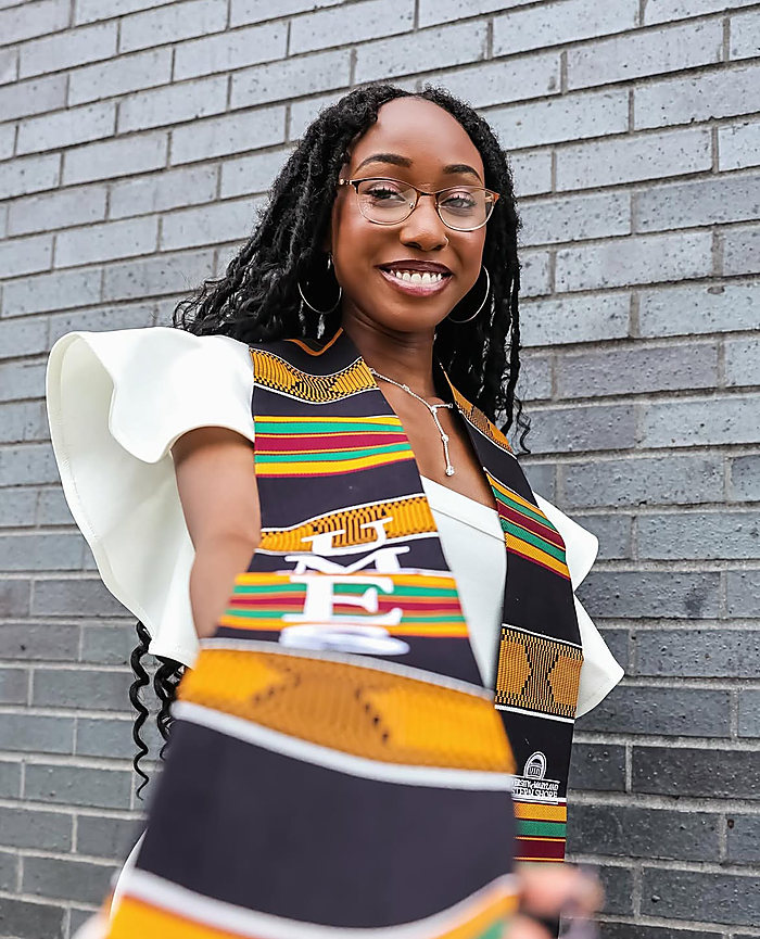 A graduate in glasses and a kente stole against a gray brick wall