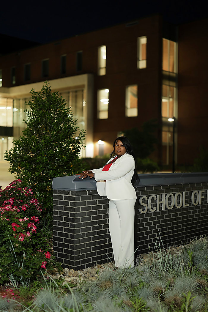 A graduate at dusk in a white suit, leaning on a brick sign outside a lit building