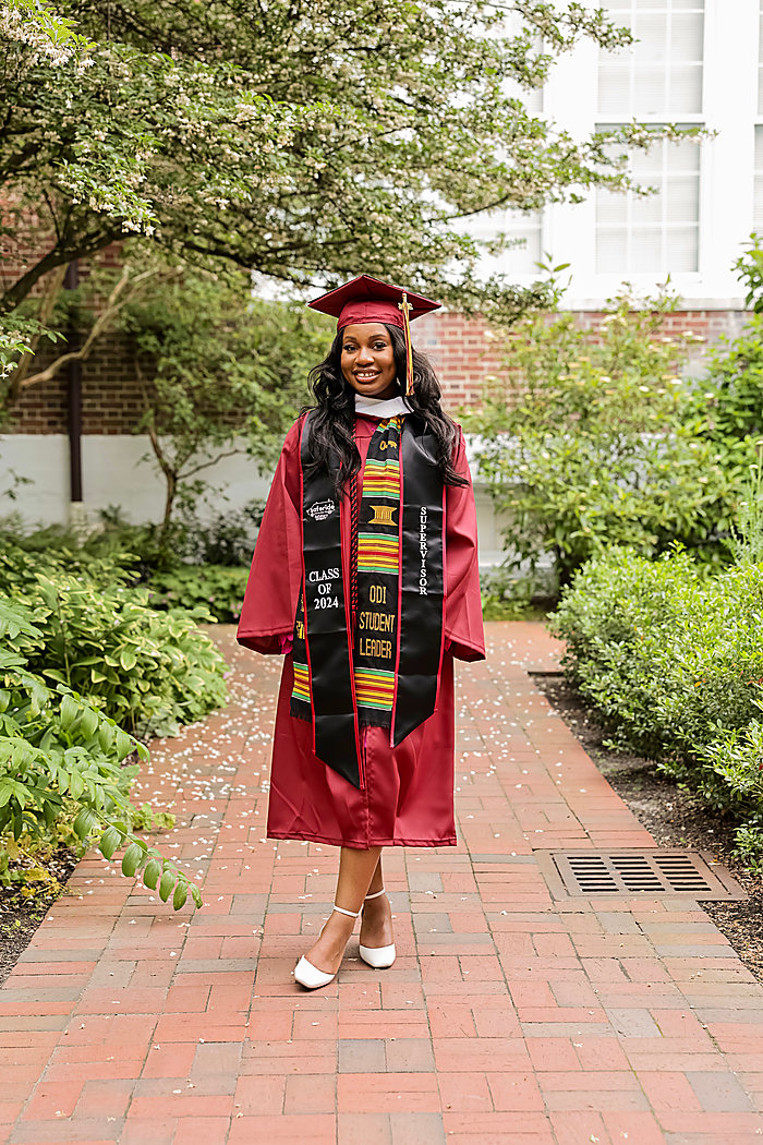 A graduate on a brick path between hedges in a red cap, gown and kente stole