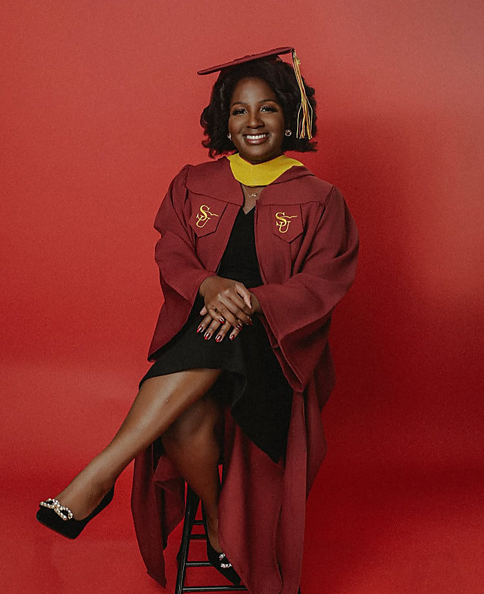 A graduate on a red backdrop, seated on a stool: a maroon gown with a gold hood