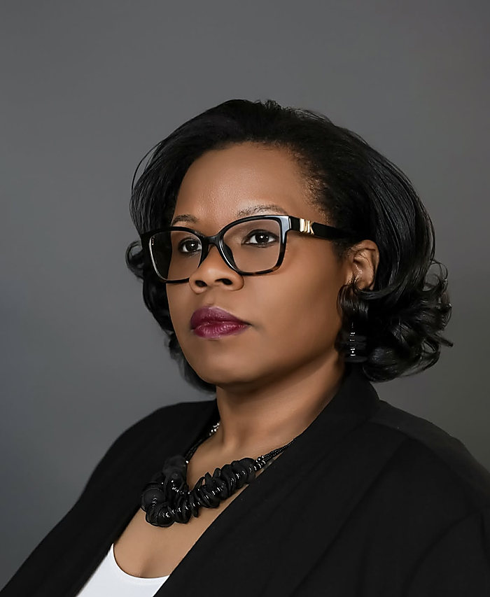 A headshot on dark gray: glasses, a black blazer and a statement necklace