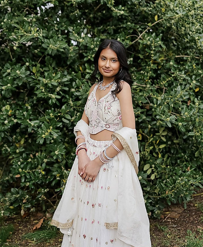 A portrait against a green hedge: a white embroidered gown, a sheer scarf and bangles