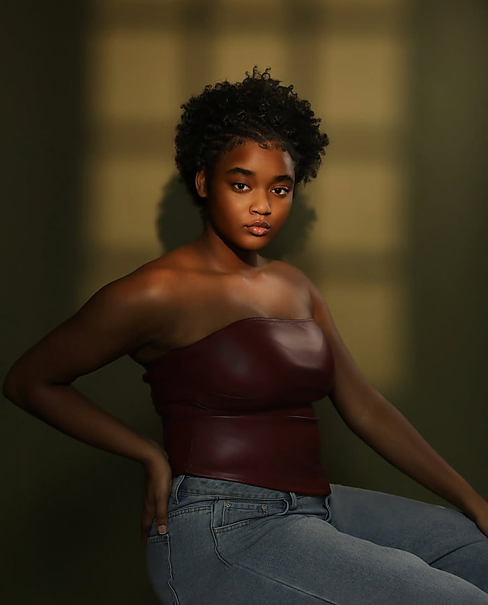 A studio portrait in low window light: an oxblood corset top and jeans, one hand on the hip