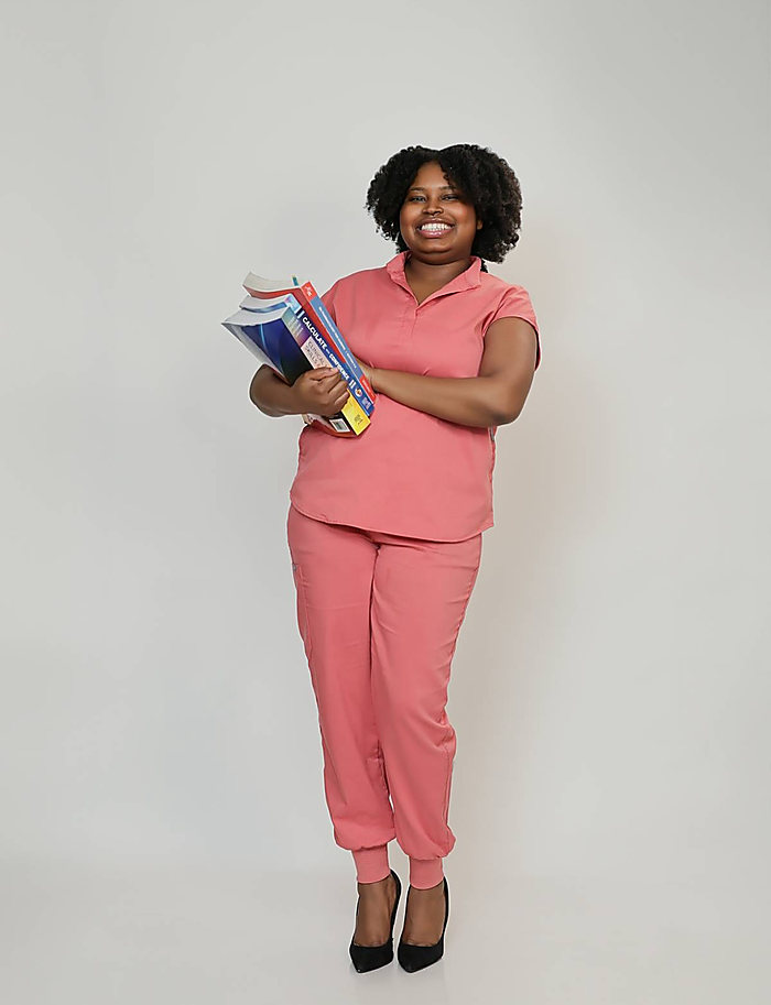 A full-length portrait on gray: pink scrubs and an armful of textbooks